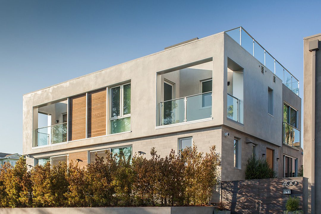 Modern two-story house with large windows, glass railings, and a beige exterior, surrounded by a hedge.