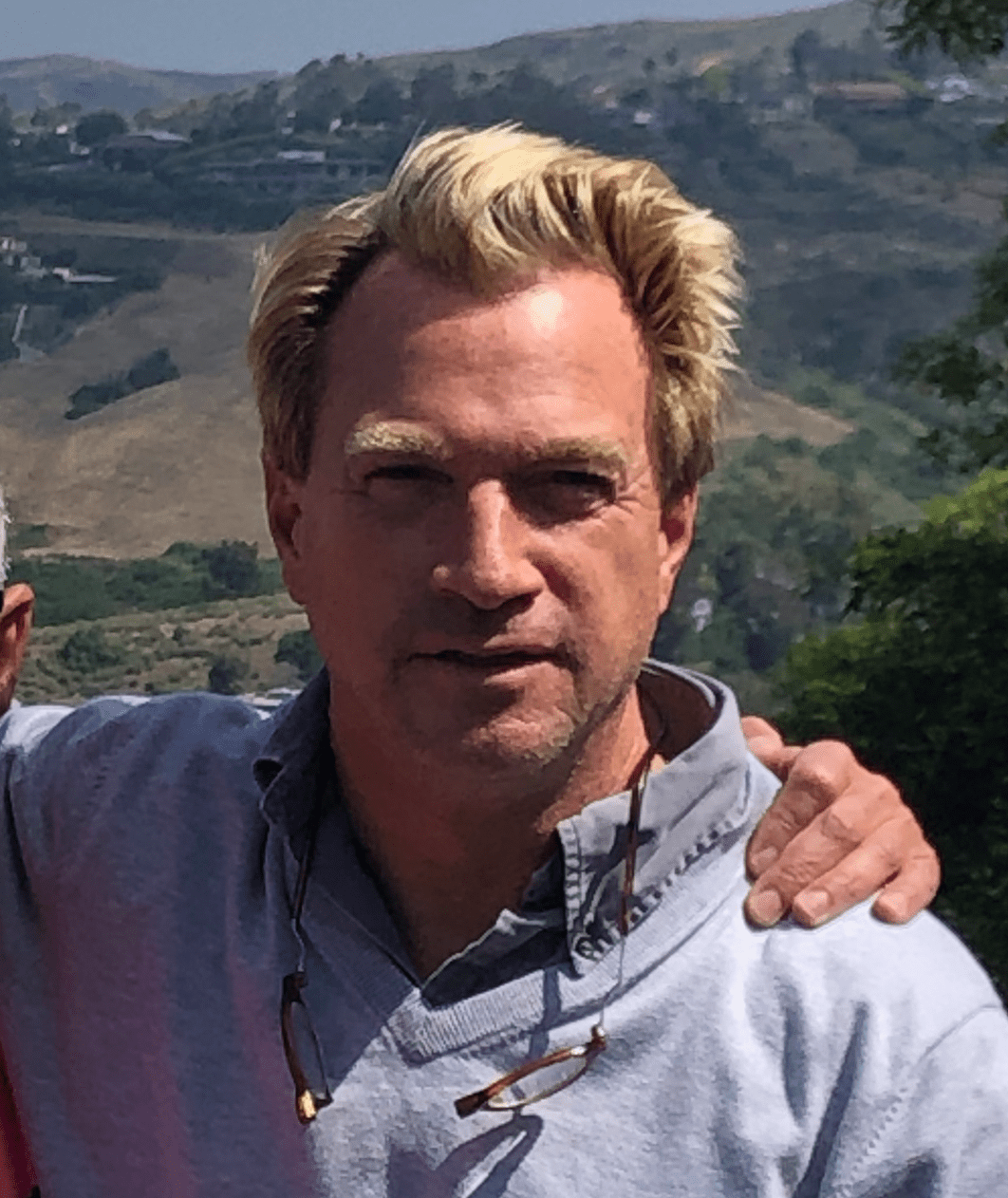 A man with blonde hair wearing a light gray collared shirt and sunglasses around his neck stands outdoors with hills in the background.