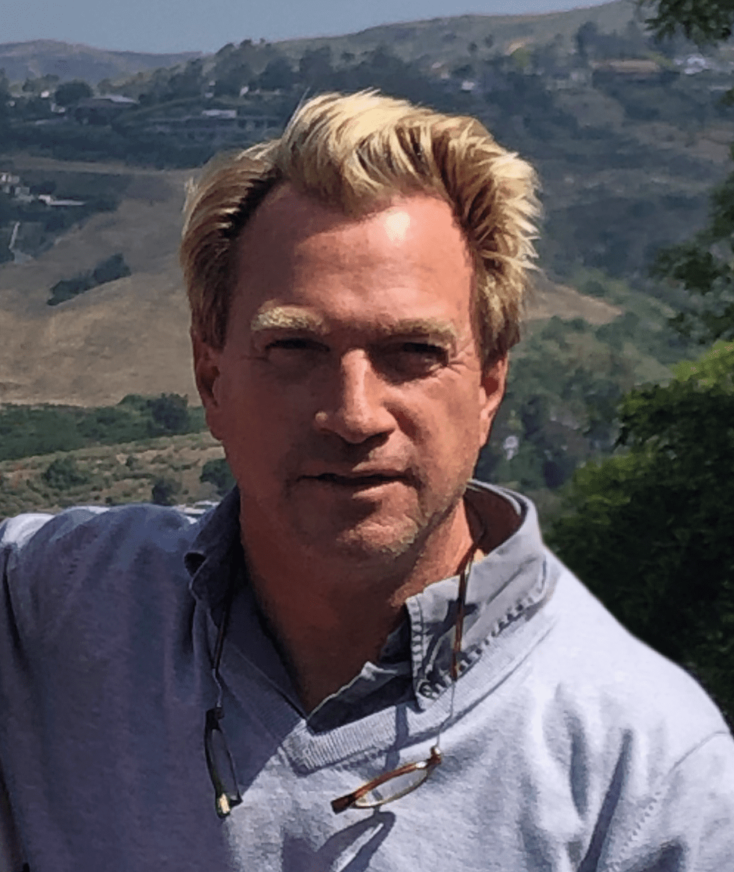 A man with blond hair wearing a light gray collared shirt and glasses hanging from his neck stands outdoors with hills in the background.